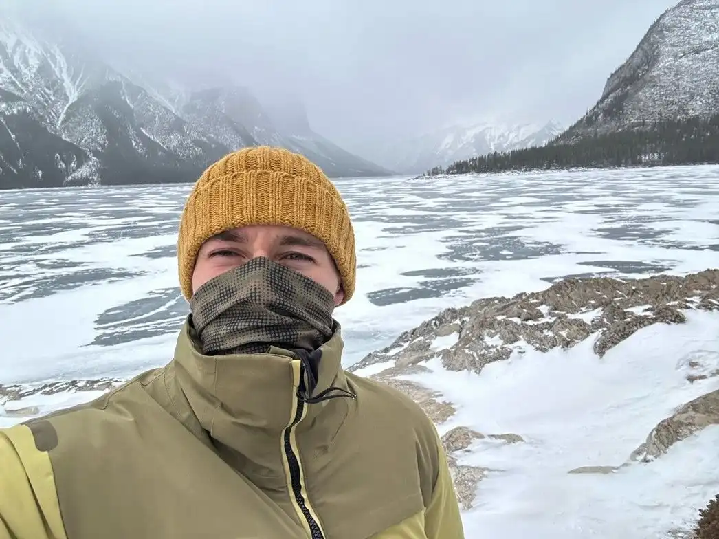 Photo showing a young man in cold weather standing in front of mountains covered with snow